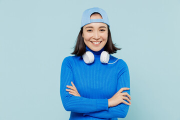 Wall Mural - Happy blithesome charismatic young woman of Asian ethnicity 20s years old wears blue shirt cap headphones around neck hold hands crossed isolated on plain pastel light blue background studio portrait.