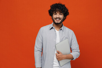 Smiling overjoyed excited happy jubilant young bearded Indian man 20s years old wears blue shirt hold use work on laptop pc computer looking camera isolated on plain orange background studio portrait.