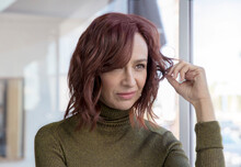 A Middle-aged Woman With Red Hair Looks Out A Window, Twirling Her Hair.