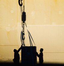 Silhouette Of Two Men Guiding Cargo Attached To Large Hook In Front Of Ships Hull