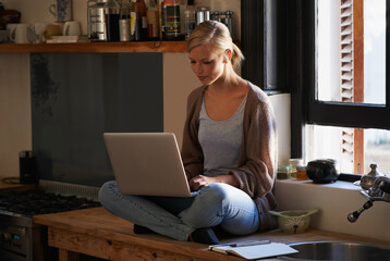 Wall Mural - Finding the right recipe online. Shot of a young woman sitting on her kitchen counter using a laptop.