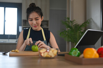 Wall Mural - Woman in apron sitting at domestic kitchen table and cutting apple on wooden board.