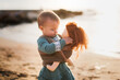 Cute european baby toddler in a green dress plays with a doll on the beach by the sea at sunset, gray background. Girl hugging waldorf soft doll, favorite toys and summer vacation kids