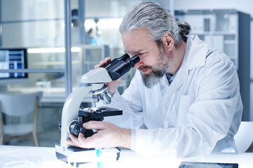  Bearded chemist in lab coat looking through the microscope and examining the virus during scientific experiment at lab
