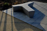 Fototapeta Przestrzenne - in the square stands a gray, white bench made of artificial stone. T or Y-shaped concrete casting. Perennial flower beds in winter covered with frost in the morning sun.