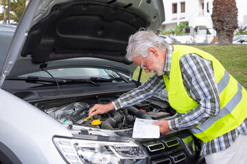 Senior driver stopped in the street for a car breakdown with the hood open wearing the yellow vest waiting for roadside assistance. Look at the instruction booklet to understand what is broken