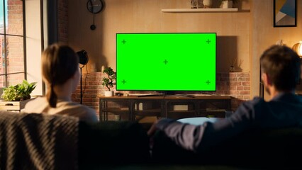 Fototapete - Authentic Couple Spending Time at Home, Sitting on a Couch and Watching TV with Green Screen Mock Up Display in Their Stylish Loft Apartment. Man and Woman Streaming Movie or Show. Shot From Back.