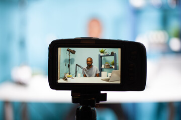 Closeup of professional digital camera on tripod recording vlogger talking at professional microphone in vlogging studio. Selective focus on back screen of dslr filming influencer sitting at desk.