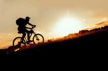 Silhouette Of A Man Riding A Bike Uphill At Sunset.