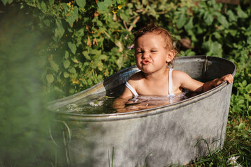  A baby smiling girl is bathing in a bathtub standing in the garden, on a hot sunny summer day
