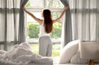 Back view of well rested young Asian woman waking up after sleeping standing up from bed and opening curtains looking at window at home in sunny morning.