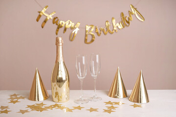 Wall Mural - Golden party caps, a bottle of champagne, glasses and an inscription happy birthday on the table. Beige festive background with copy space.