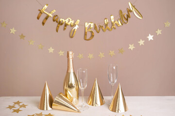Wall Mural - Golden party caps, a bottle of champagne, glasses and an inscription happy birthday on the table. Beige festive background with copy space.