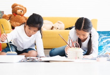 Adorable little kid and her sister lying on floor having fun using brushes and colorful paint drawing on paper. Playful childhood siblings spend leisure time together playing in living room as artist.