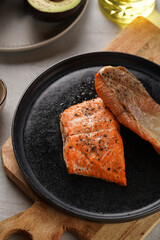 Two grilled fish steaks - red salmon on black round plate on wooden board, olive oil, avocado on grey background