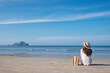 © Farknot Architect - Rear view image of a young woman with hat and bag sitting on the beach with blue sky background