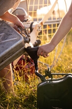 Close-up Of Male Hands Pouring Gasoline From One Canister To Another Using A Plastic Funnel. Fuel For Paragliding On A Sunny, Warm Evening.