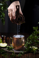 Wall Mural - Woman hand pouring Green Apple Irish Mule cocktail cocktail from shaker in copper mug surrounded by ingredients and bar tools