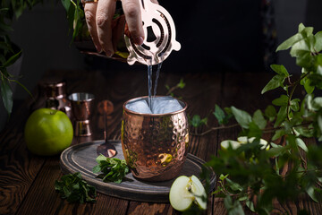 Wall Mural - Woman hand pouring Green Apple Irish Mule cocktail cocktail from shaker in copper mug surrounded by ingredients and bar tools