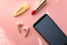 Mobile Phone, Pink Wireless Earphones, Red Lipstick And Pearl Hair Clip On Pastel Pink Background. Flat Lay.