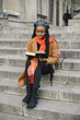 © prostooleh - Young black woman in a coat sitting on a stairs and reading a book