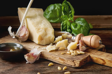 Italian parmesan cheese with knife fresh green basil, olive oil, pine nuts and garlic. Ingredients for the pesto. Dark wood background