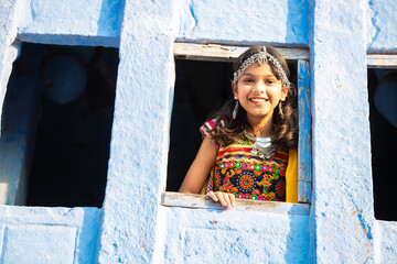 Happy little indian girl kid wearing traditional colorful rajasthani outfit and jewelery looking out of window.