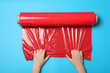 Woman unrolling red plastic stretch wrap on light blue background, top view