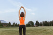 Back view of fit sportswoman in bright orange activewear stretching arms and warming up while training on field in summer 