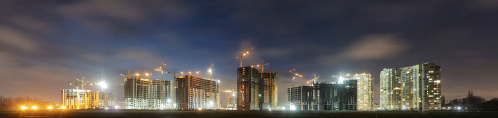 View of a large construction site with buildings under construction and multi-storey residential homes.Tower cranes in action on night sky background. Panoramic view.