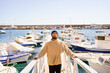 © javiemebravo - young man 30-40 years old standing on a harbor gangway with his hands resting, he is wearing a turtleneck and a wool hat and is looking at the camera