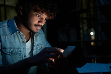 Young indian man using mobile apps technology on cell phone late at night searching information in internet, reading news, making digital payments in applications on smartphone, communicating online.