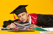 © master1305 - Portrait of young man, student in graduation cap and gown falling asleep on books isolated over yellow studio background