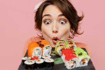 Wall Mural - Close up shocked surprised amazed impressed happy young woman 20s hold in hand look at makizushi sushi roll served on black plate traditional japanese food isolated on plain pastel pink background