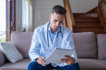 Portrait of handsome young male doctor in white lab coat with stethoscope looking at reports in medical office. Caucasian male healthcare worker in uniform analyzing written report or test results 