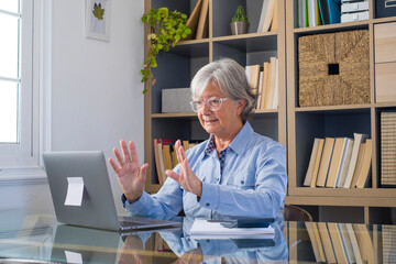 Senior businesswoman video chatting online using laptop with adhesive note at home office in front of book shelf. Elderly caucasian woman gesturing while working online or watching media content 