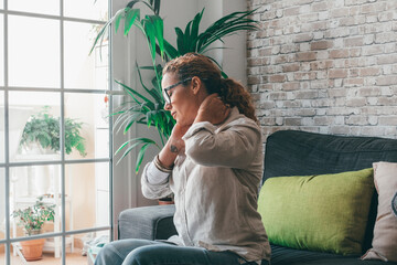 Exhausted young woman suffering from strong neck ache while sitting on sofa in the living room of house, Caucasian female sitting on couch touching and massaging tensed neck muscles feeling discomfort