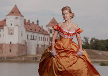 A Young Woman In An Old Orange Dress Walks On The Shore Of The Lake Near The Castle