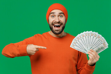 Wall Mural - Young surprised excited fun happy caucasian man 20s wear orange sweatshirt hat holding point index finger on fan of cash money in dollar banknotes isolated on plain green background studio portrait