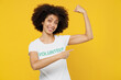 © ViDi Studio - Young woman of African American ethnicity in white volunteer t-shirt point on biceps muscles demonstrate strength power isolated on plain yellow background Voluntary free work assistance help concept