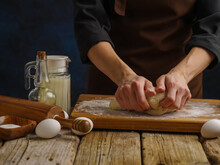 Close-up. The Chef Kneads The Dough On A Wooden Cutting Board, Dark Blue Background. Eggs, Flour, Milk, Butter, Kitchen Utensils On A Wooden Table. Recipes For Dough Products.