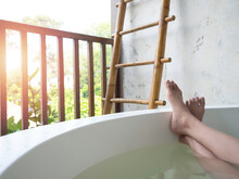 Women's Feet In A White Bathtub With Full Clear And Clean Water Near The Bamboo Ladder And Wooden Fence And Leaves On Outdoor Nature View.