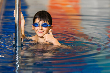 A little boy in swimming goggles holds on to the pool railing and shows OK. Swimming school. Copy space.