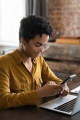 Pensive positive young African gadget addict user woman using cellphone at workplace, sitting at laptop, chatting online, browsing Internet, shopping with ecommerce app. Communication gadgets