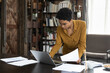 © fizkes - Focused serious young Black businesswoman holding, checking documents, working at laptop computer. African worker, professional using computer in home office, doing paperwork job, standing at table