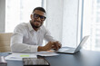 Portrait of smiling millennial African American confident male manager employee sitting at table with computer, posing in modern office, motivated young businessman leader worker looking at camera.