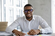 © fizkes - Front view smiling young handsome African American male executive manager leader in eyeglasses wearing wireless earphones holding negotiations video call online meeting with colleagues in office.