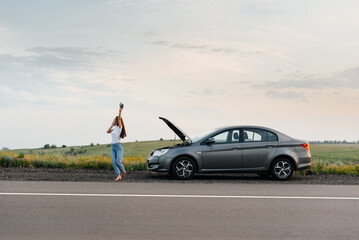 Wall Mural - A young girl stands near a broken car in the middle of the highway during sunset and tries to call for help on the phone and start the car. Waiting for help. Car service. Car breakdown on the road.