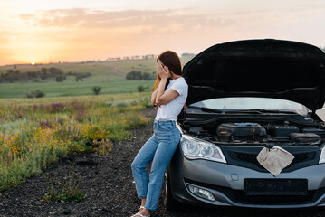 Wall Mural - A frustrated young girl stands near a broken-down car in the middle of the highway during sunset. Breakdown and repair of the car. Waiting for help. Car service. Car breakdown on road.