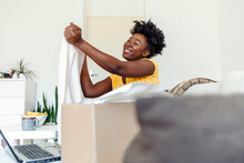 Portrait Of Curious Cheerful Black Woman Received Package, Unpacking Cardboard Box, Looking Inside, Sitting On The Sofa In Living Room At Her Apartment. Happy With Online Purchase.